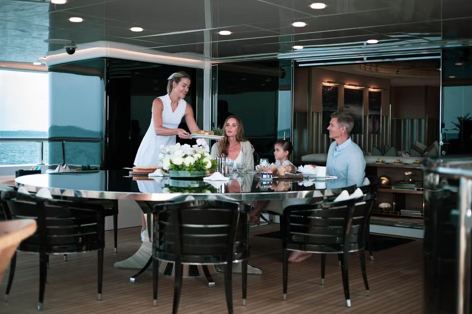 Crew member serving gourmet meal to family dining in yacht interior with panoramic sea views